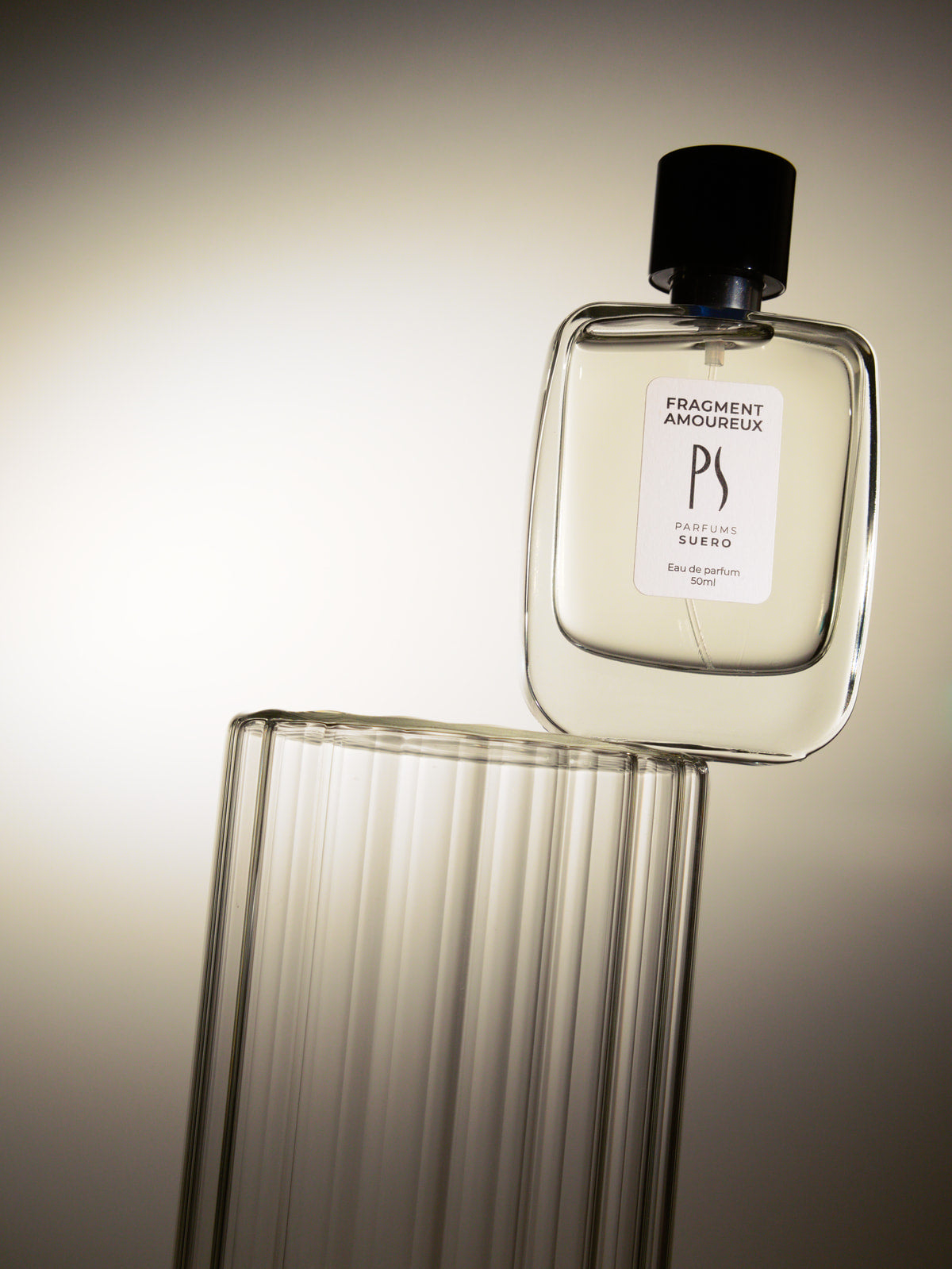 Flacon penché du parfum Fragment Amoureux de la marque PARFUMS SUERO le flacon est posé sur une colonne en verre il est en équilibre et transparent suggérant la légèreté 