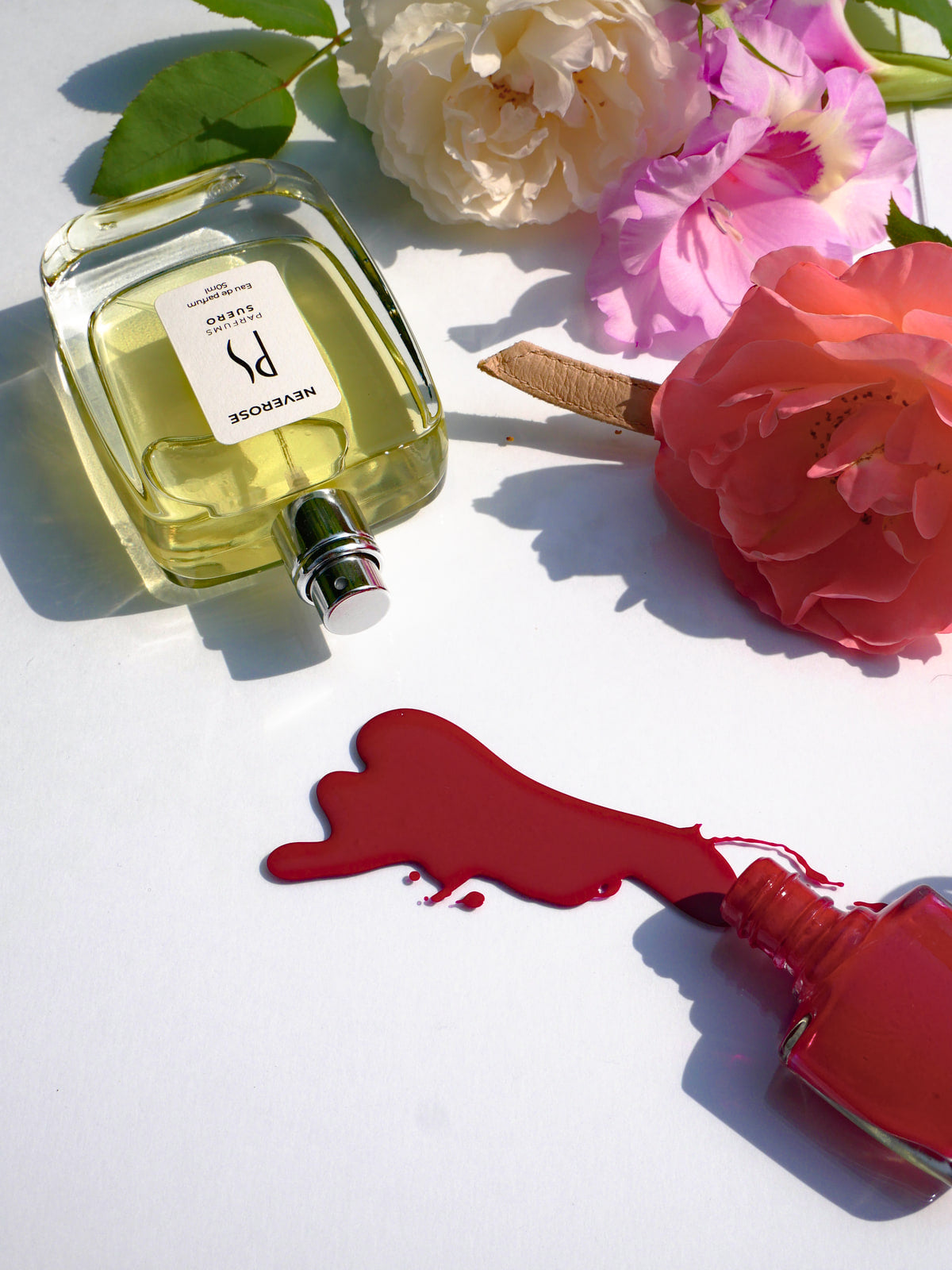 un flacon du parfum Neverose de la marque PARFUMS SUERO est renversé au soleil au milieu de fleurs un flacon de vernis à ongle rouge est aussi renversé