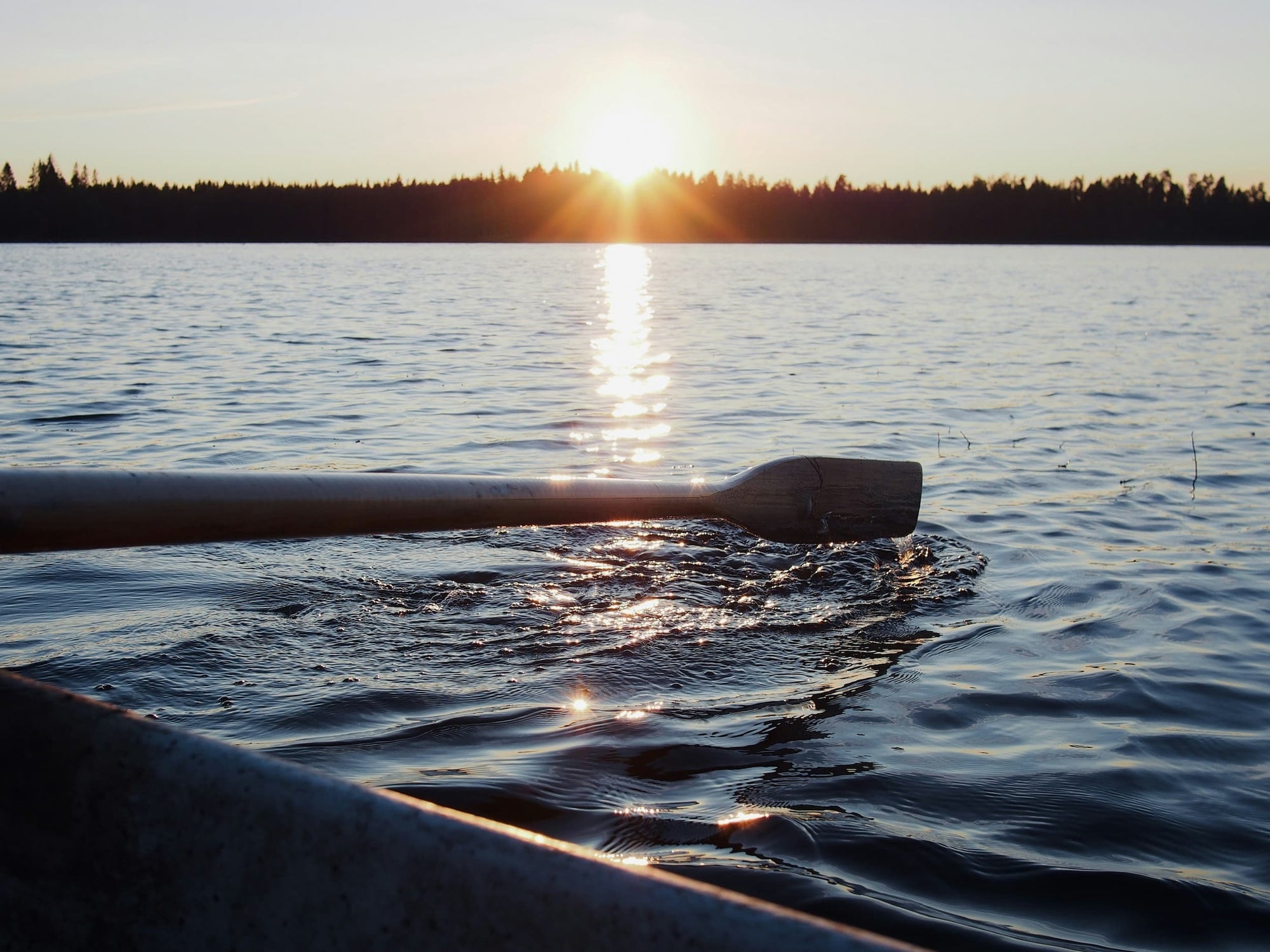 Pagaie posée sur l’eau calme d’un lac au coucher de soleil, avec le reflet du soleil créant une traînée lumineuse à la surface de l’eau