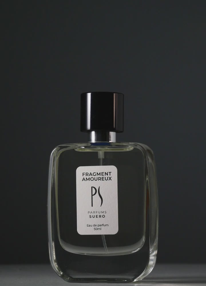 un flacon de Fragment Amoureux de la marque PARFUMS SUERO apparait doucement dans une lumière progressive qui laisse apparaitre un fond sur lequel se reflète des végétaux
